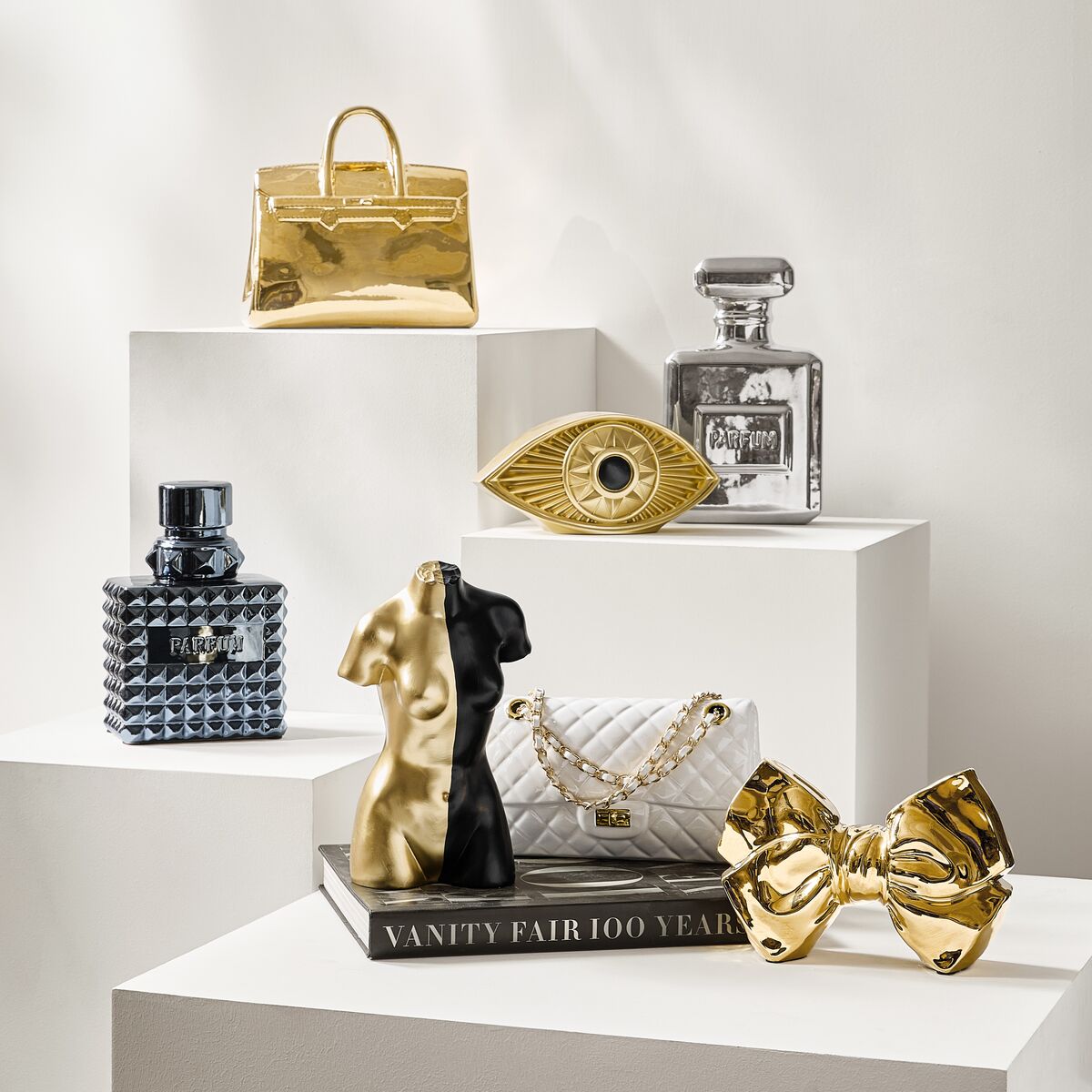 Chic luxury gift display featuring a gold handbag sculpture, perfume bottle decanter, gold bow accent, and black figural bookends on white plinths.