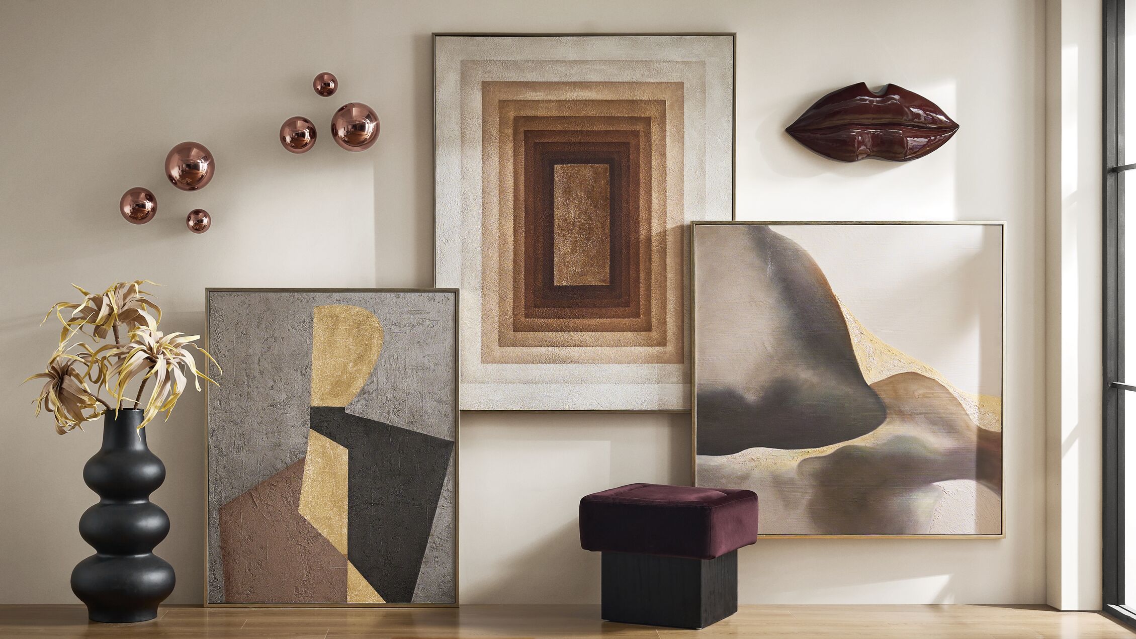 Gallery wall displaying textured neutral framed abstract art, bronze chrome sphere wall accents, a sculptural maroon lip wall art piece, paired with a black totem floor vase and a plum velvet ottoman.