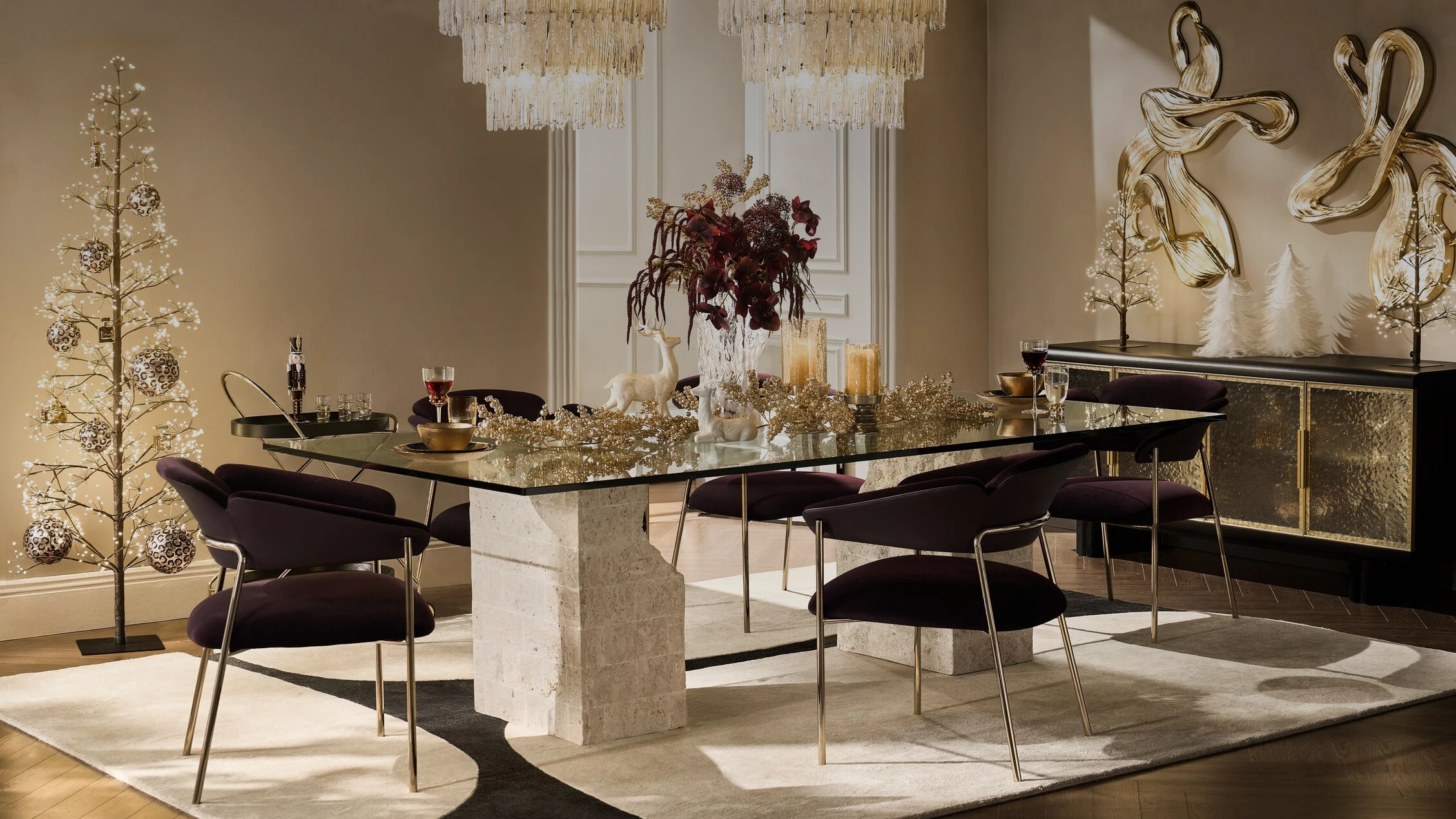 Champagne Velvet Dining Room featuring a glass top dining table with travertine stone pedestals, deep eggplant velvet dining chairs, golden holiday table garland, and sparkling crystal chandeliers.
