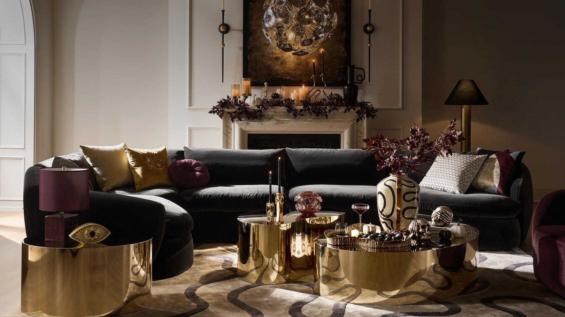 Modern Mystique Living Room featuring a dark velvet sectional sofa, reflective brass coffee tables, moody candlelit fireplace mantel, and dramatic globe chandelier.