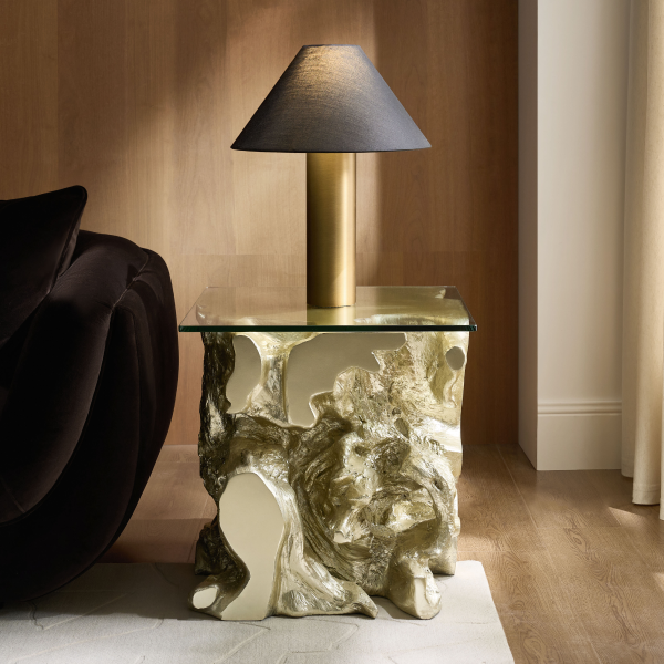 Modern side table featuring a gold sculptural molten-look base with a glass tabletop, topped with a brass table lamp with a conical shade.