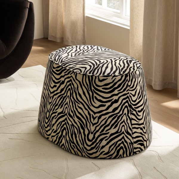 Oval ottoman stool upholstered in black and white zebra pattern fabric, placed on a light neutral area rug near a sunlit window.