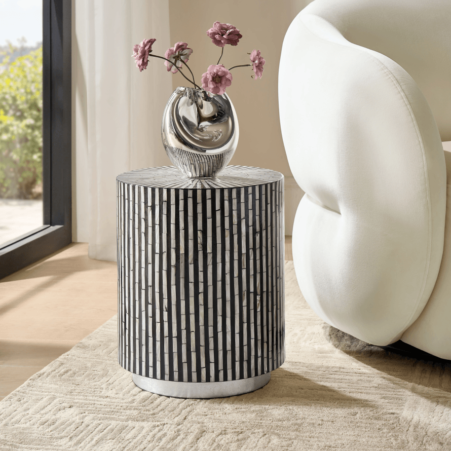 Black and white striped cylindrical end table topped with a silver vase and pink flowers next to a white curved sofa.