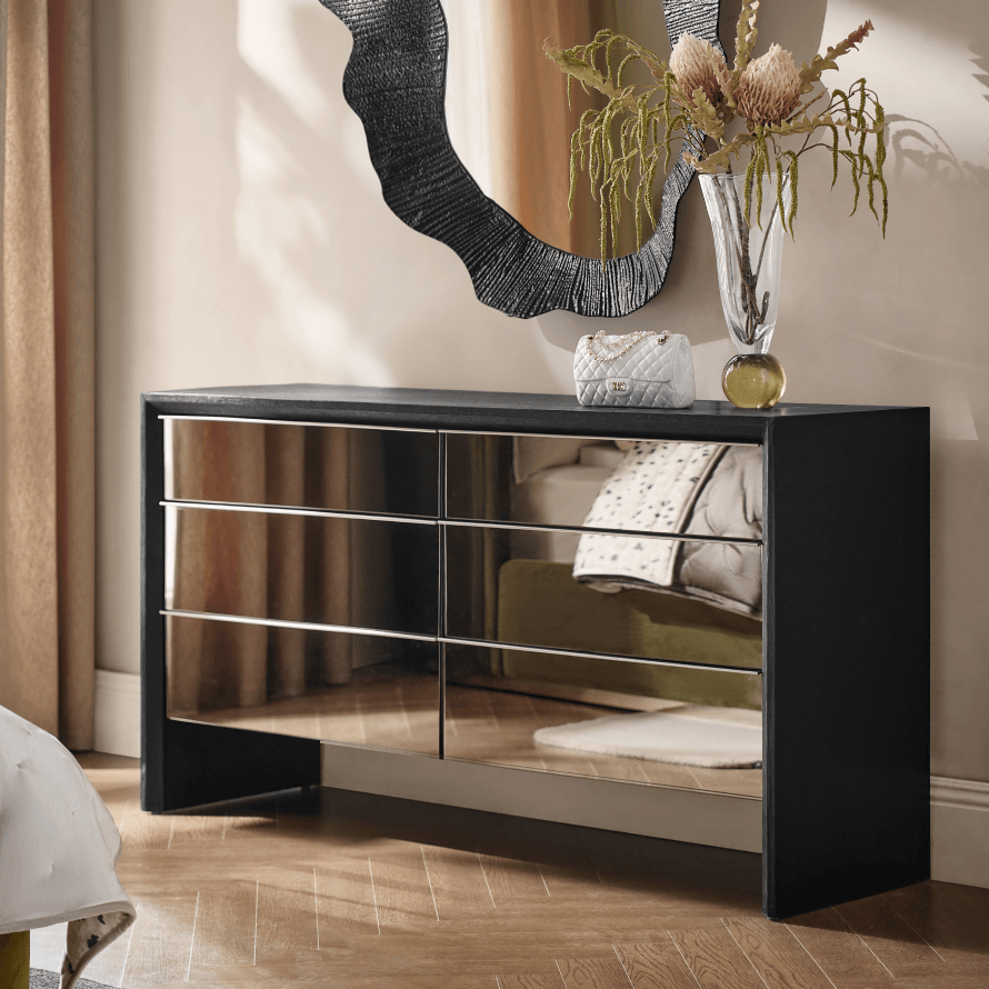 Contemporary black six-drawer dresser with reflective mirrored front panels placed under an organic wavy wall mirror.