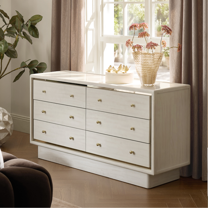 Modern ivory double dresser with brass hardware and subtle texture, styled next to a sunlit window with curtains and a indoor tree.