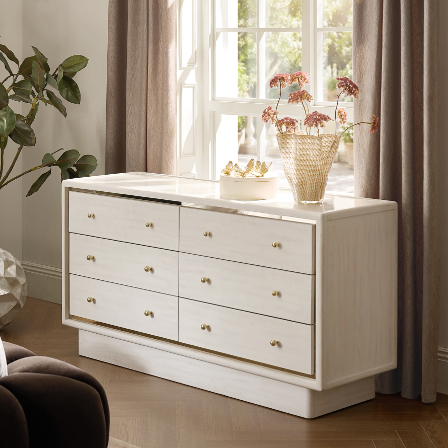 Modern six-drawer cream lacquered double dresser with gold hardware and mirrored trim styled with dried florals and decorative accents.