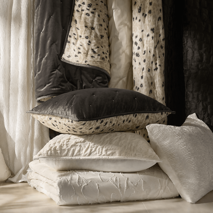 Stacked luxury bedding collection featuring dark charcoal velvet pillows, leopard print accents, textured white comforters, and crinkled satin shams.