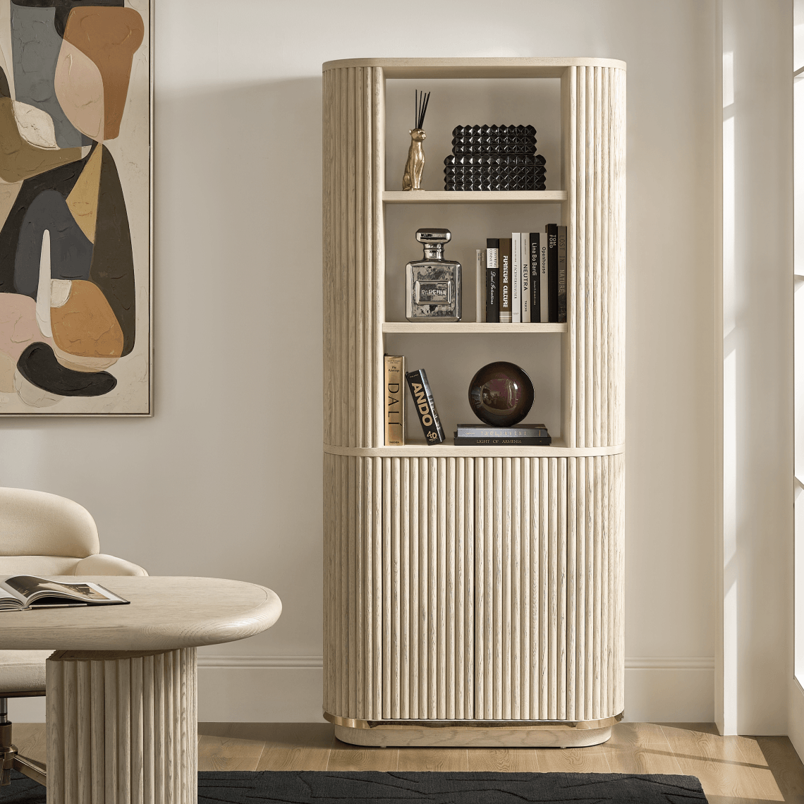 Tall fluted cream bookcase styled with books, decorative boxes, and sculptures next to abstract wall art.