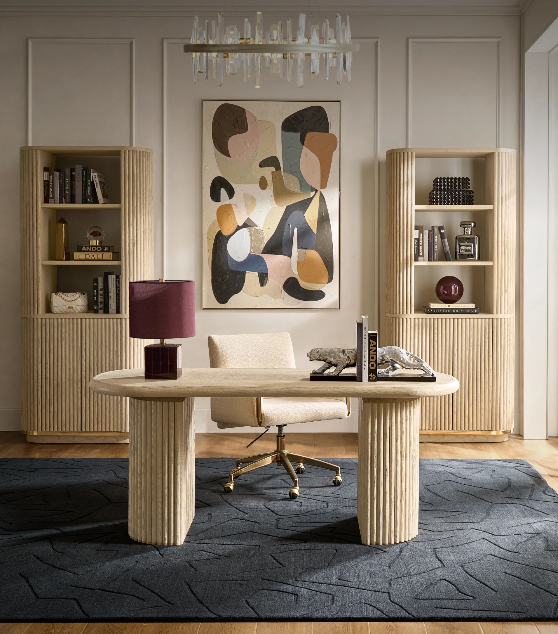 Modern home office featuring a fluted wooden executive desk, cream swivel desk chair, pair of tall fluted bookcases, and abstract painting.