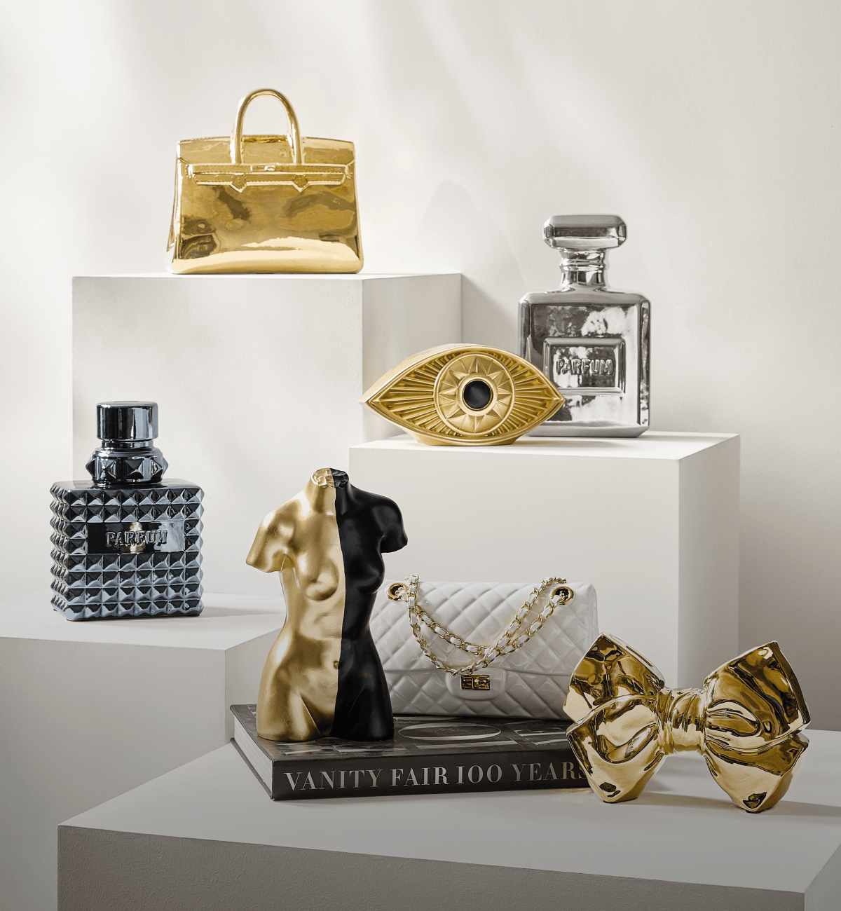 Curated luxury decorative objects including a gold handbag sculpture, perfume bottles, evil eye accent, and split gold and black torso figure on display pedestals.