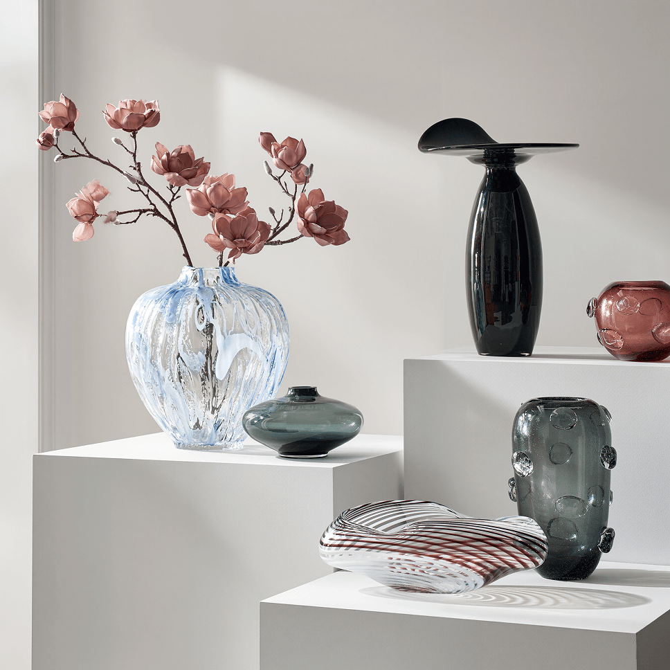 Modern sculptural glass and ceramic vases with floral branches displayed on white gallery pedestals.