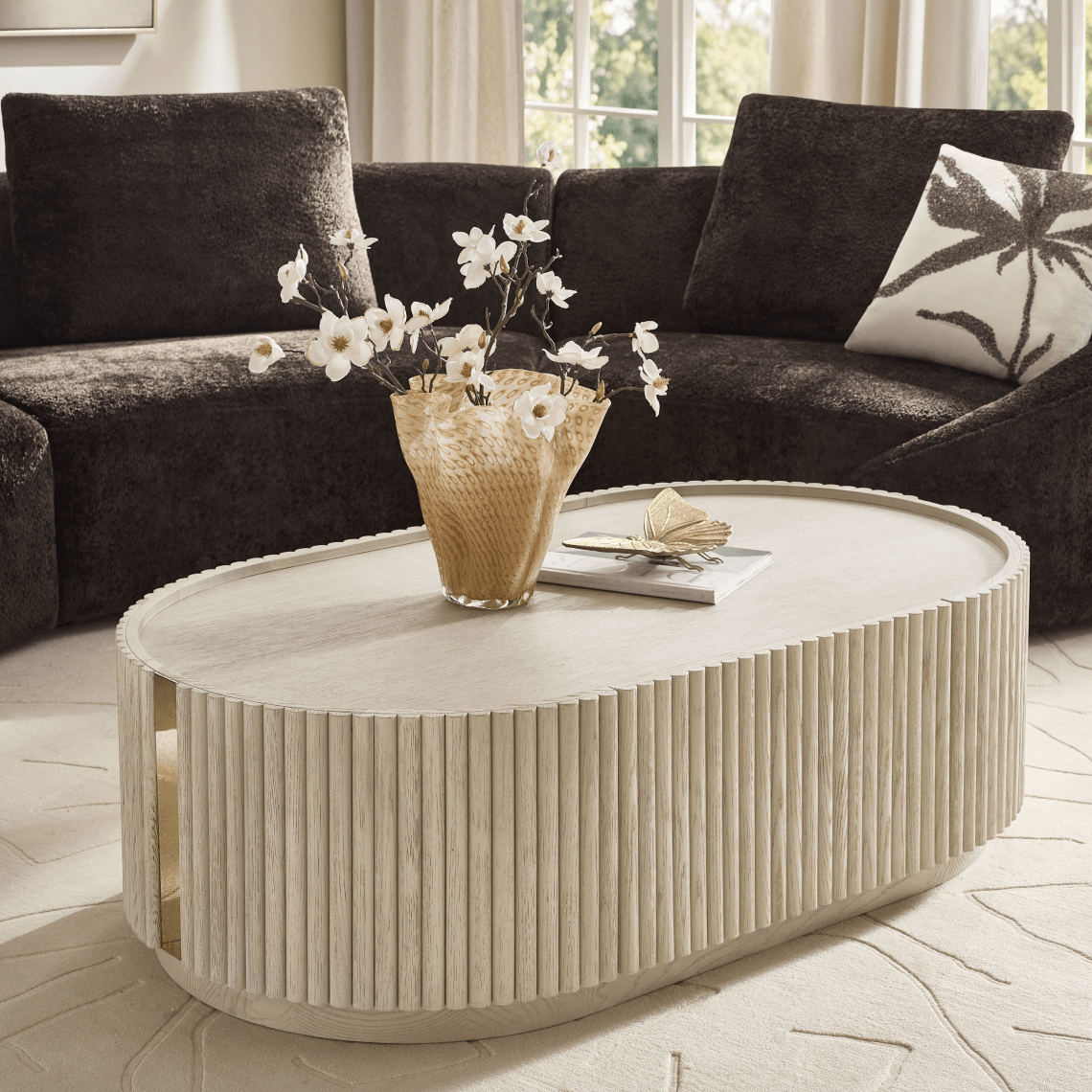 Oval fluted cream coffee table styled with a vase of white florals, set in front of a dark brown plush sectional sofa.