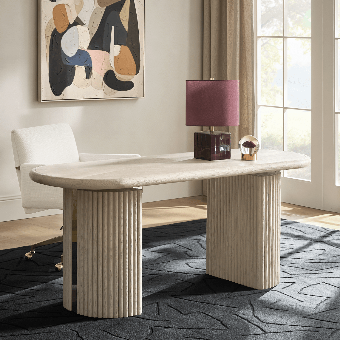 Oval executive desk with fluted pillar legs topped with a dark lamp, paired with a white desk chair and dark patterned rug.
