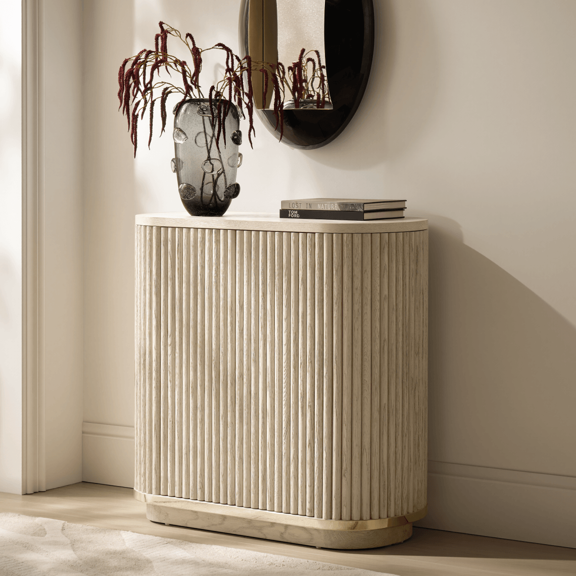 Tall fluted cream buffet cabinet featuring brass base trim, topped with a smoked glass vase and dark oval mirror.