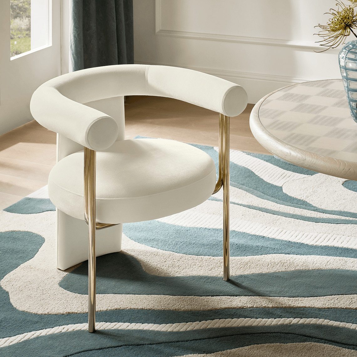 Modern ivory upholstered curved barrel dining chair with slim brass legs positioned on a blue and neutral wavy area rug.