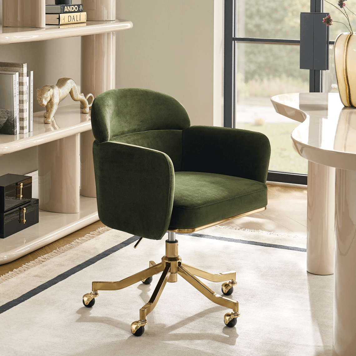 Forest green velvet swivel desk chair with brass caster base positioned in a light-filled modern home office.