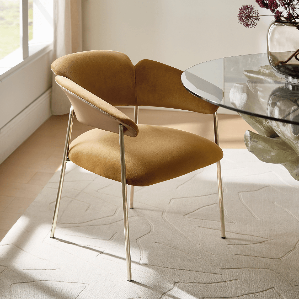 Camel velvet curved dining chair with sleek brass legs positioned near a glass-top dining table.