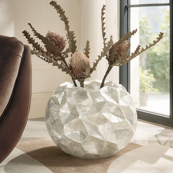 Large white geometric faceted ceramic vase filled with dried floral branches, set on a floor near a window.