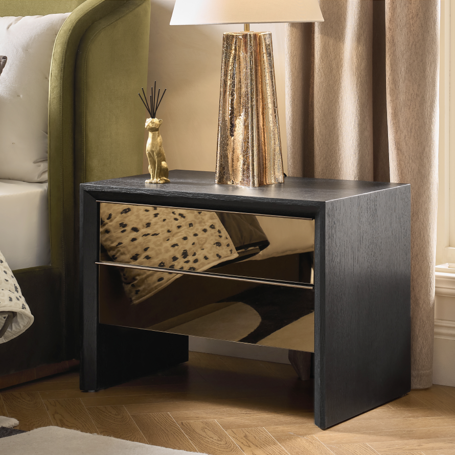 Dark oak nightstand with mirrored drawer fronts, topped with a tall metallic brass table lamp next to an upholstered bed.