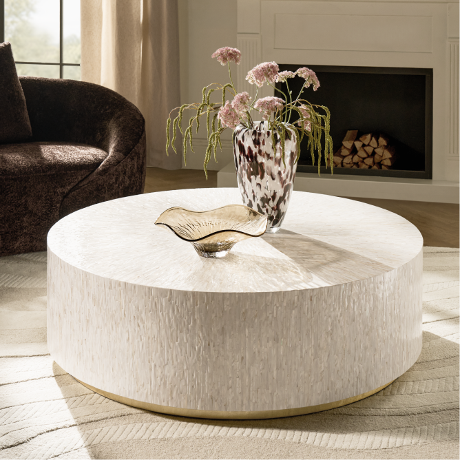 Round ivory drum coffee table with brass base and textured finish, styled with a speckled glass vase in a modern living room.