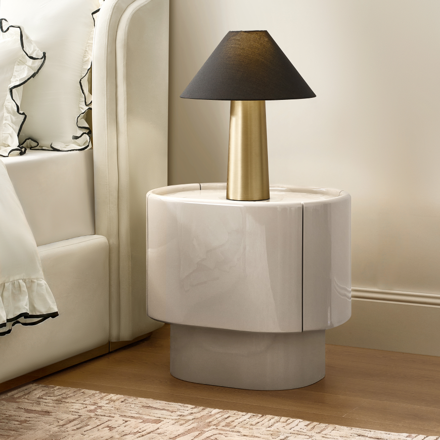 Modern curved high-gloss cream lacquered nightstand topped with a brass cone table lamp beside an upholstered shelter bed.
