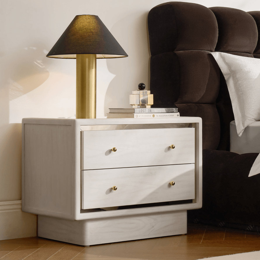 Off-white two-drawer nightstand with brass knobs topped with a cylinder lamp, set next to a dark tufted bed frame.
