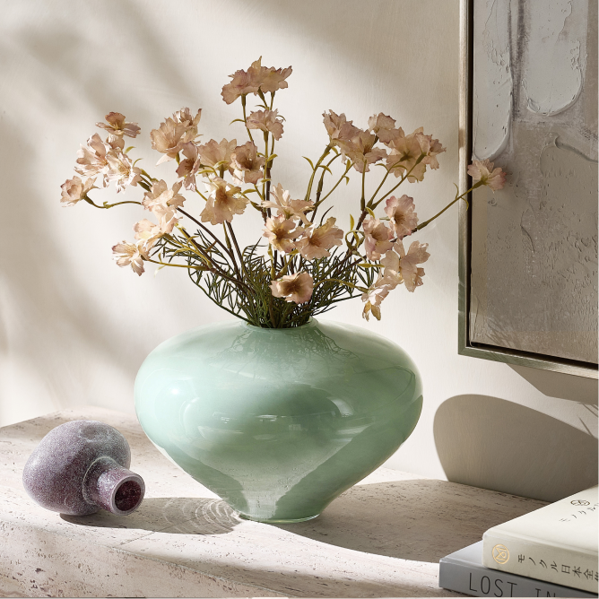 Light mint green ceramic vase holding soft pink faux florals next to a small purple accent vessel on a stone tabletop.