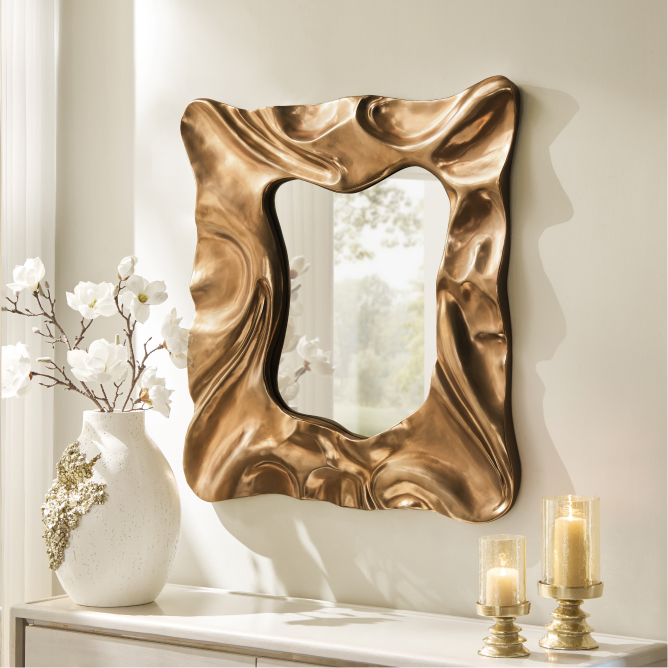 Sculptural organic gold wall mirror mounted above a credenza with pillar candle holders and a decorative white floral vase.