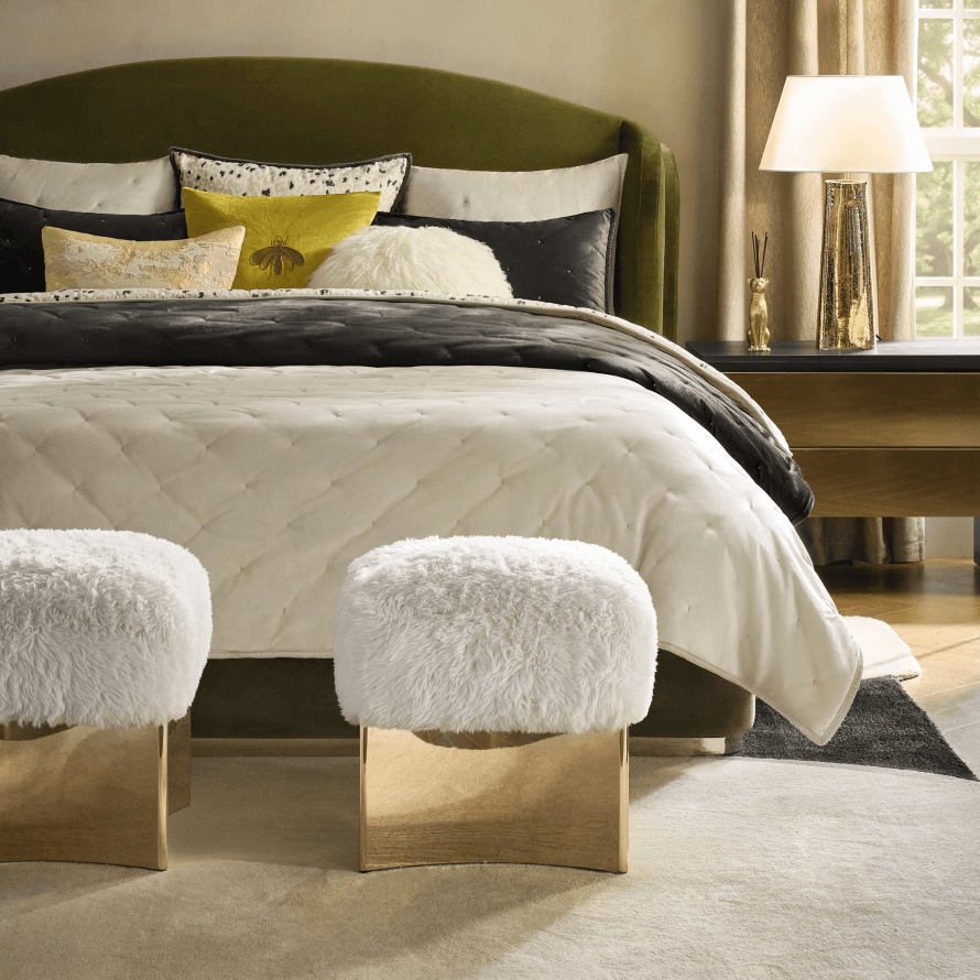 Olive green upholstered bed styled with layered neutral bedding, decorative throw pillows, and two white fuzzy ottomans with brass bases.