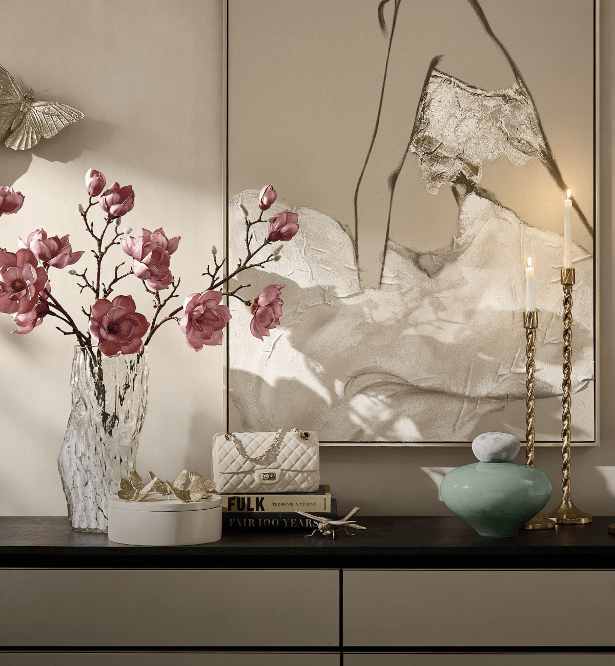 Elegant console table setting featuring pink magnolia flowers in a clear vase, brass candlesticks, a green decorative jar, and a large framed artwork.