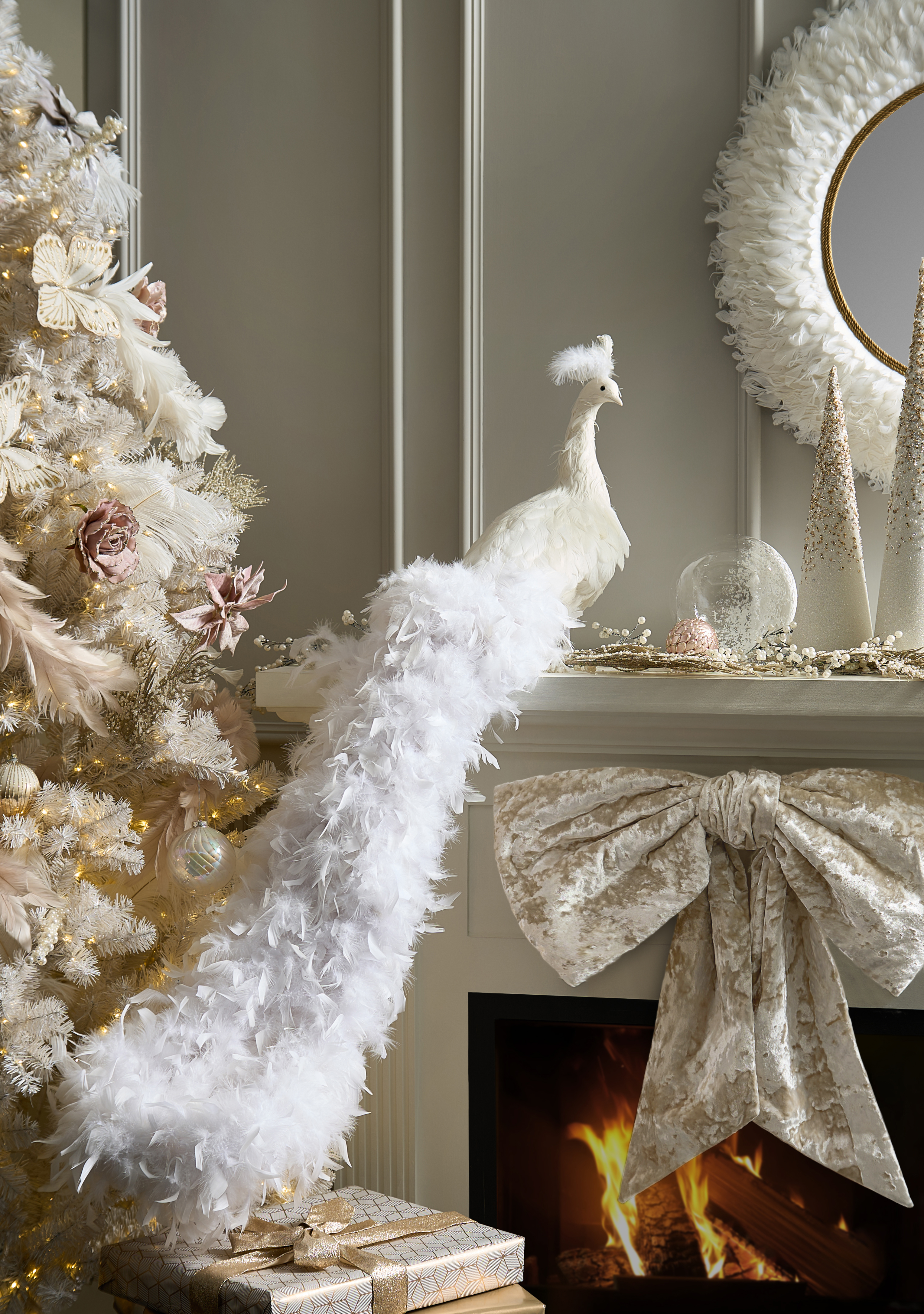 Elegant white peacock with a long feathered tail perched on a Christmas mantel with a feather wreath, glittered trees, and a large velvet bow above a fireplace.