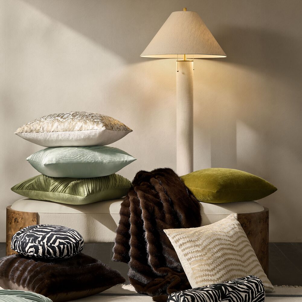 Stacked and styled decorative throw pillows in sage green, metallic gold, velvet olive, and zebra print, paired with a dark faux fur throw blanket on a bench under a tall modern floor lamp.
