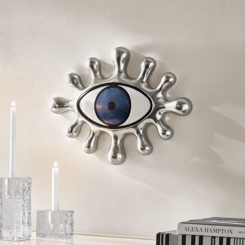 Sculpted silver metallic eye wall art mounted above crystal candleholders on a white gallery wall.