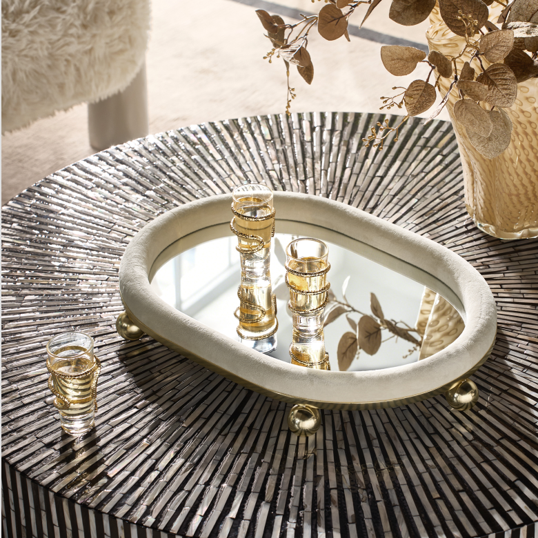 Oval mirrored decorative tray with brass ball feet styled on a metallic radial tabletop with glassware.