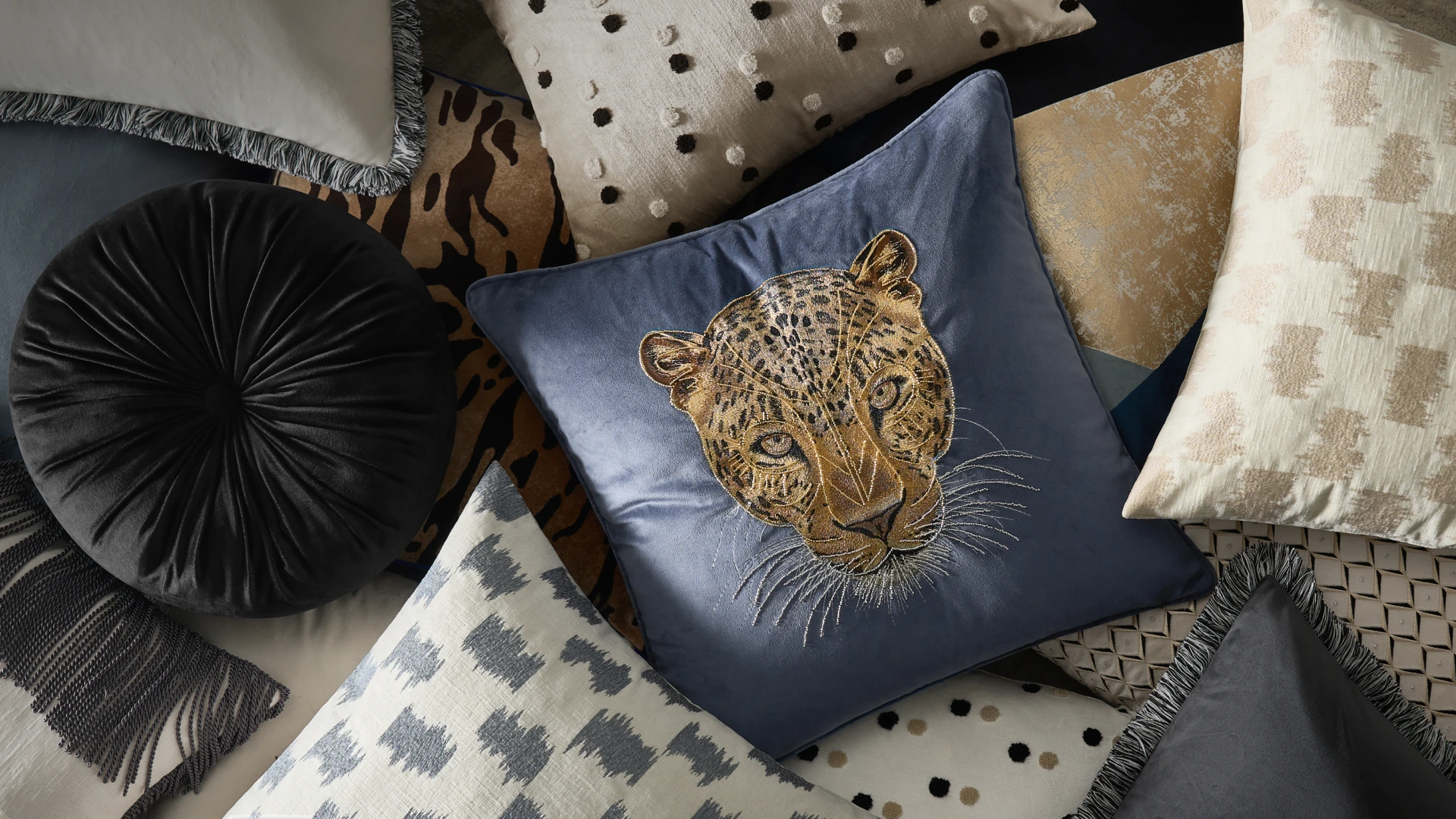 Layered collection of luxury textured accent pillows and throws featuring an embroidered velvet leopard cushion, round pleated pillow, and fringe details.
