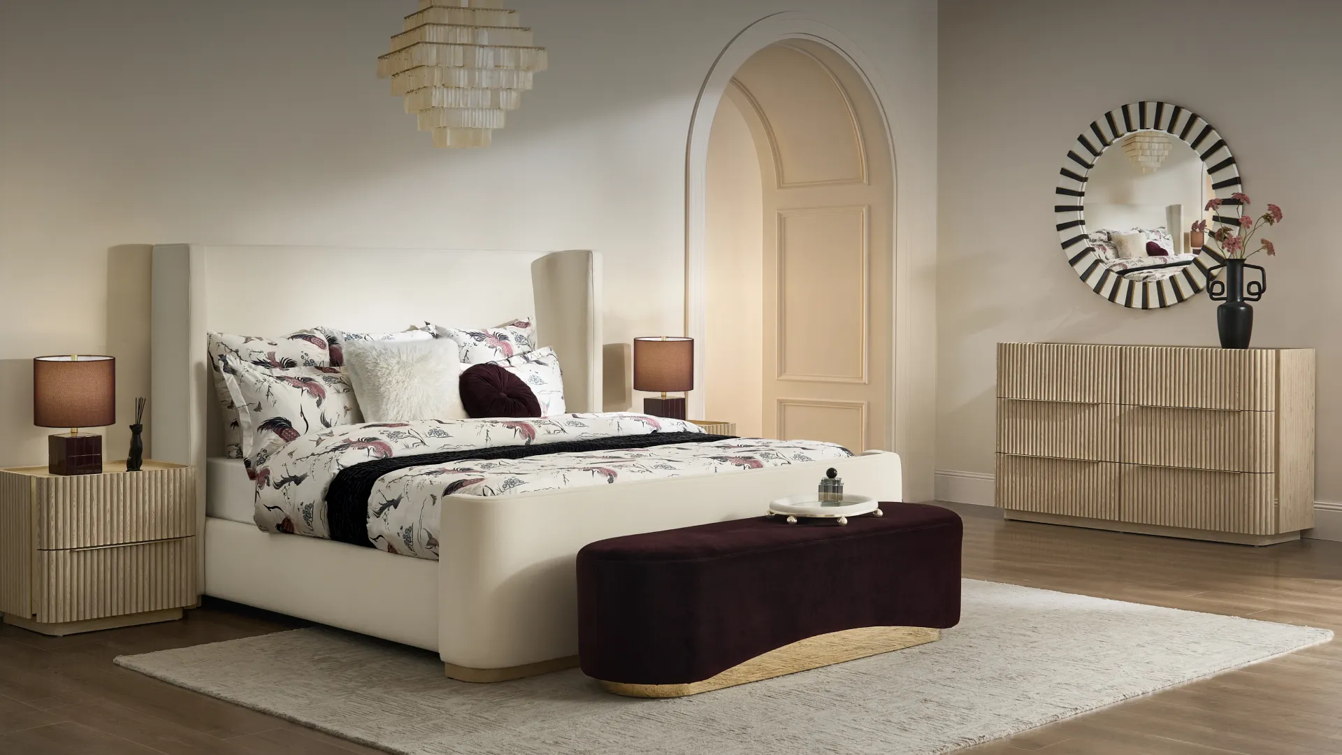 Modern luxurious bedroom featuring a cream upholstered wingback bed with floral bedding, a curved burgundy velvet bench, fluted wood nightstands, and a sunburst accent mirror.