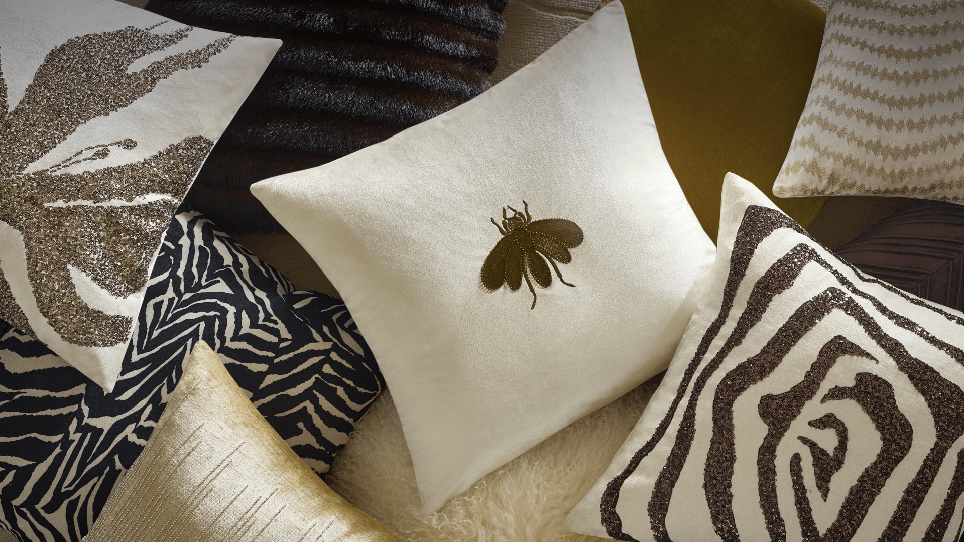Flat lay display of luxury decorative throw pillows featuring sequined metallic leaf designs, zebra prints, embroidered bee motifs, and rich velvet fabrics.