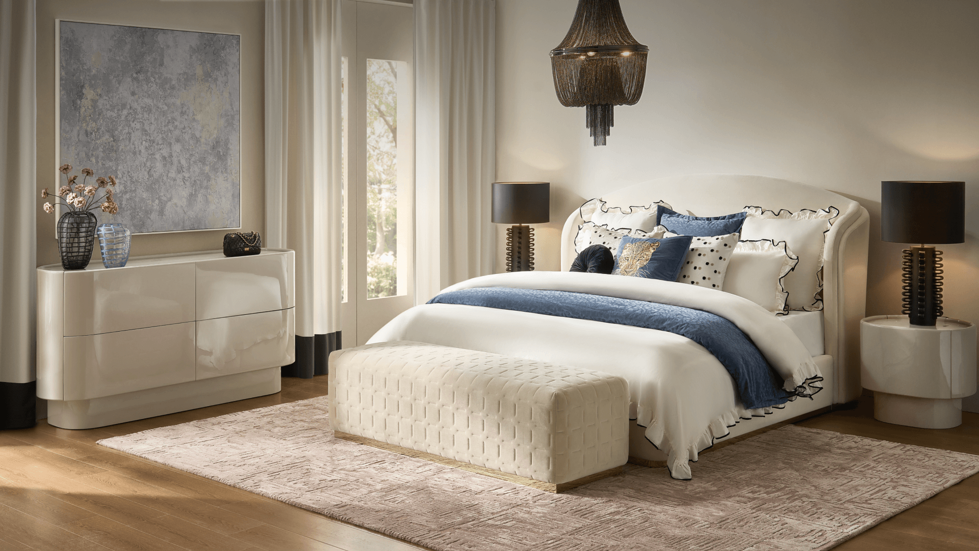 Elegant bedroom featuring an ivory upholstered shelter bed styled with blue accent bedding, woven end-of-bed bench, high-gloss cream dresser, and modern black table lamps.