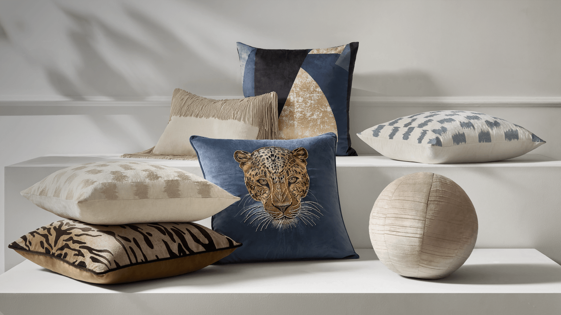 Artistic display of luxury accent pillows featuring embroidered blue leopard velvet cushion, tiger print pillow, textured sphere pillow, geometric patterns, and fringe details on gallery steps.