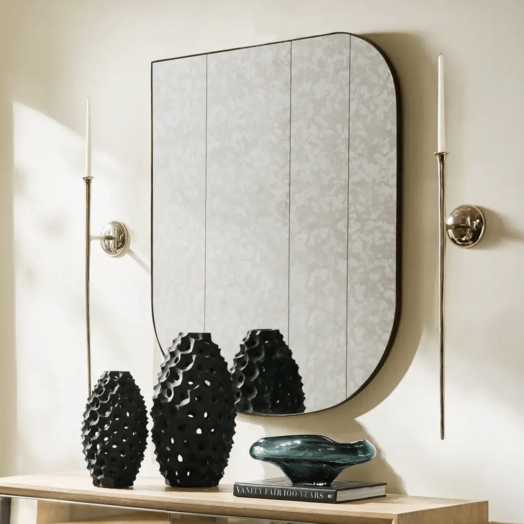 Modern vignette showcasing a Z Gallerie mirror with a unique rounded-edge rectangular shape and subtly patterned glass panels. Flanked by sleek silver wall sconces, the mirror hangs above a light wood console table styled with sculptural black vases, a smoky glass bowl, and stacked coffee table books, creating a refined and artful display.