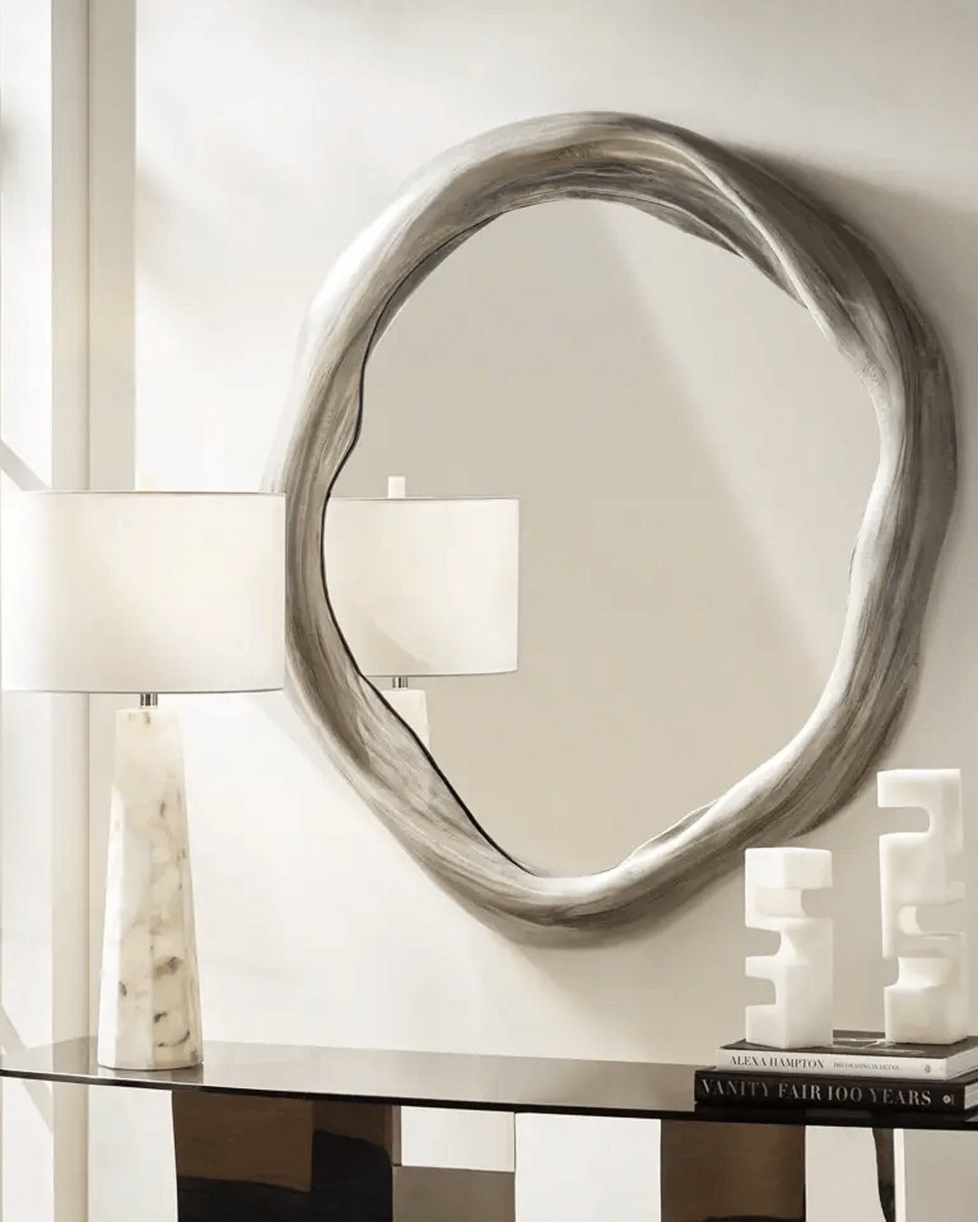 Refined modern entryway featuring a Z Gallerie mirror with an organic, sculptural silver frame mounted on a neutral wall. Styled with a marble table lamp, abstract white sculptures, and a mirrored console table topped with stacked design books, the scene blends soft curves with polished materials for an elevated, artistic look.