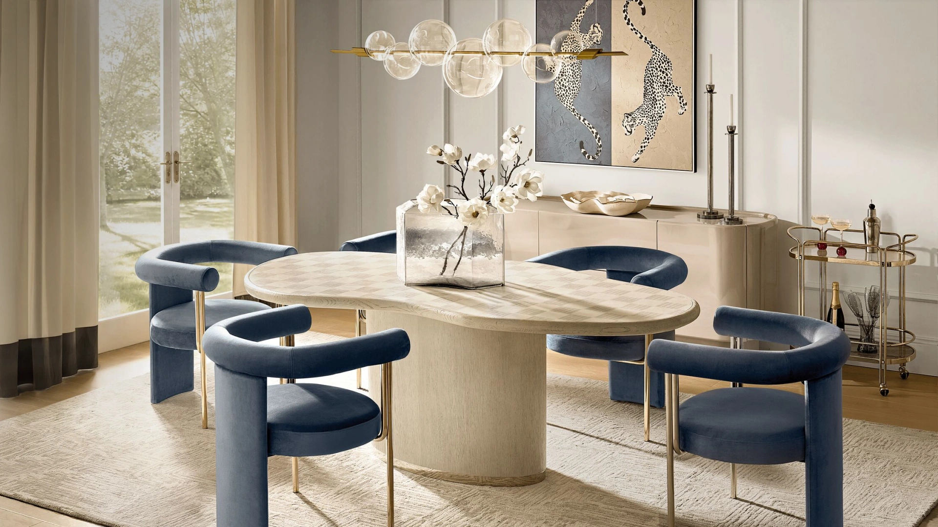 Elegant dining room featuring an organic oval light wood dining table, curved deep blue velvet dining chairs, a linear bubble glass chandelier, and rolling brass bar cart.