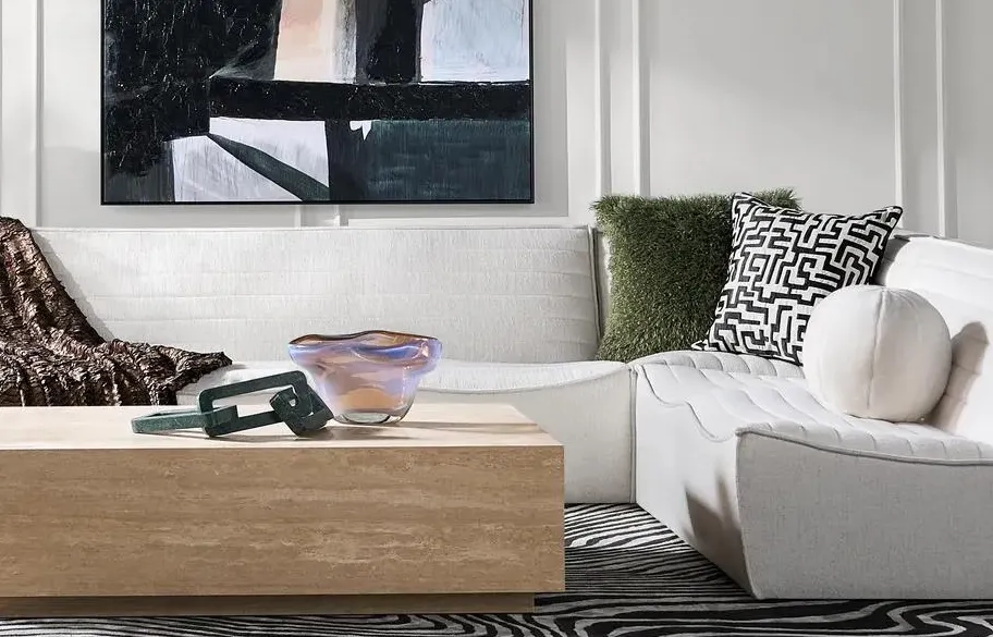 Contemporary neutral living room with a white sectional, green and black accent pillows, abstract art, and a tinted glass bowl on a wood coffee table.