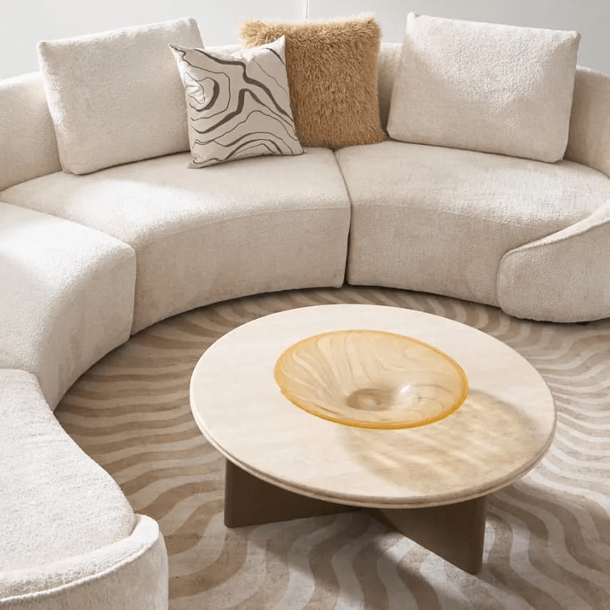 Modern curved bouclé sectional in neutral tones with textured pillows, round coffee table, amber glass bowl, and wavy beige area rug.