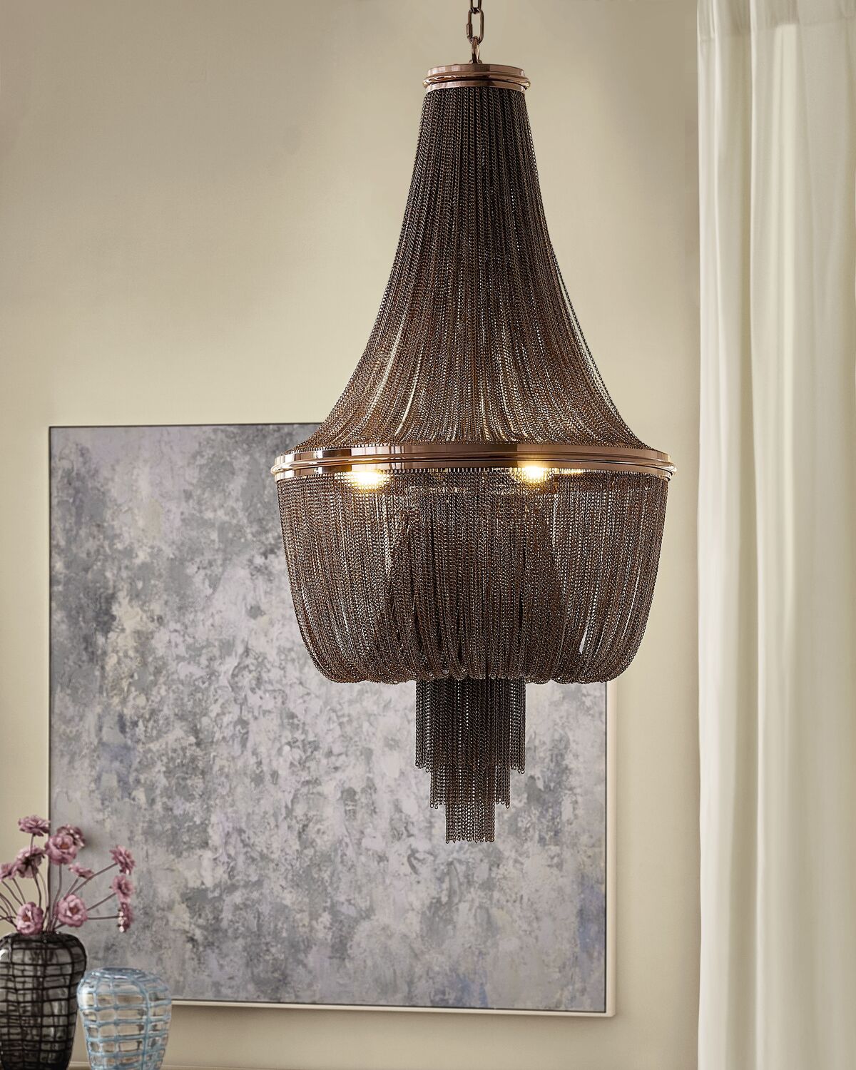 Elegant statement empire chandelier draped with cascading dark metallic chain fringe over a polished brass ring, hung in front of abstract wall art.