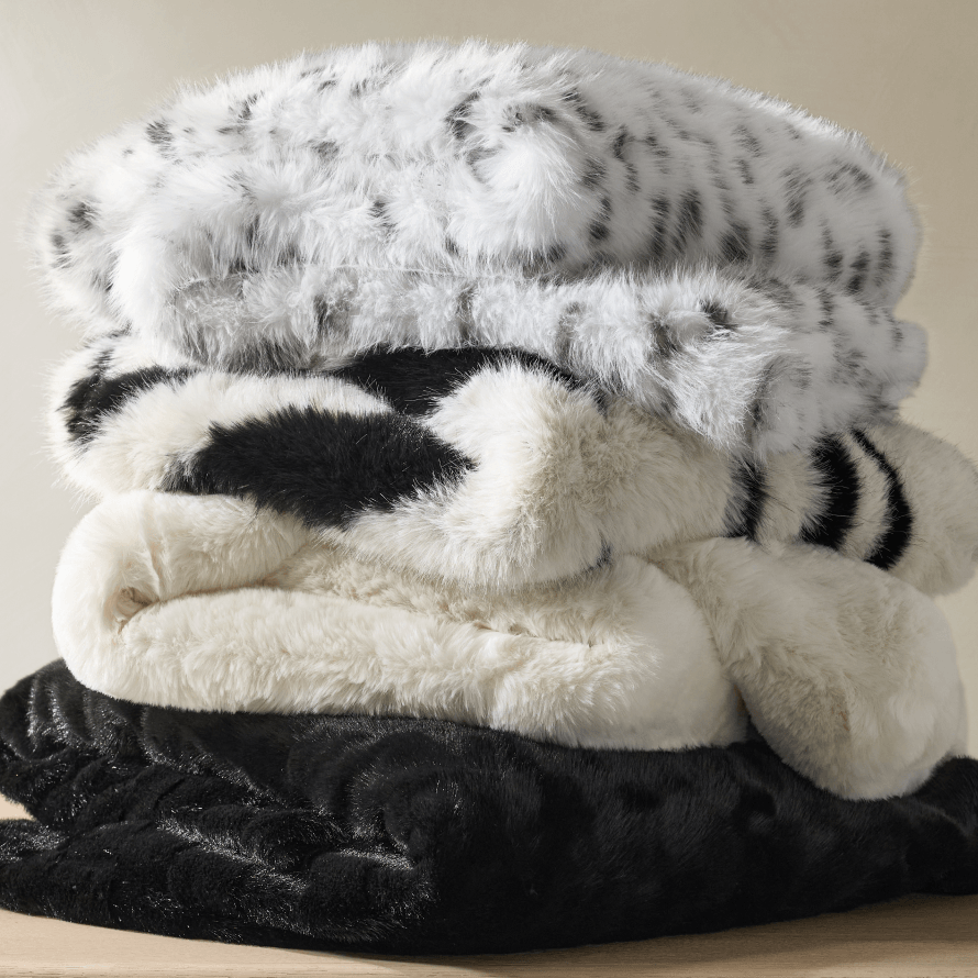 Stack of luxury faux fur and textured throw blankets featuring snow leopard print, zebra pattern, plush cream fur, and black textured velvet.
