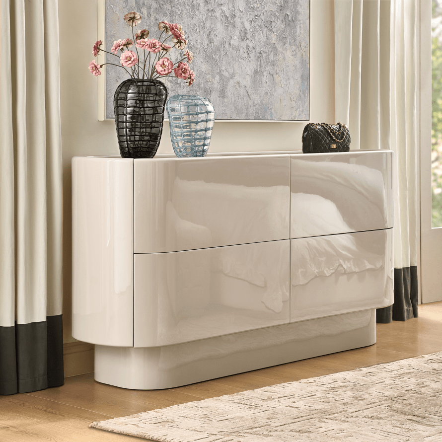 Modern curved high-gloss cream lacquered dresser styled with dark textured glass vases, pink blossoms, and framed canvas art.