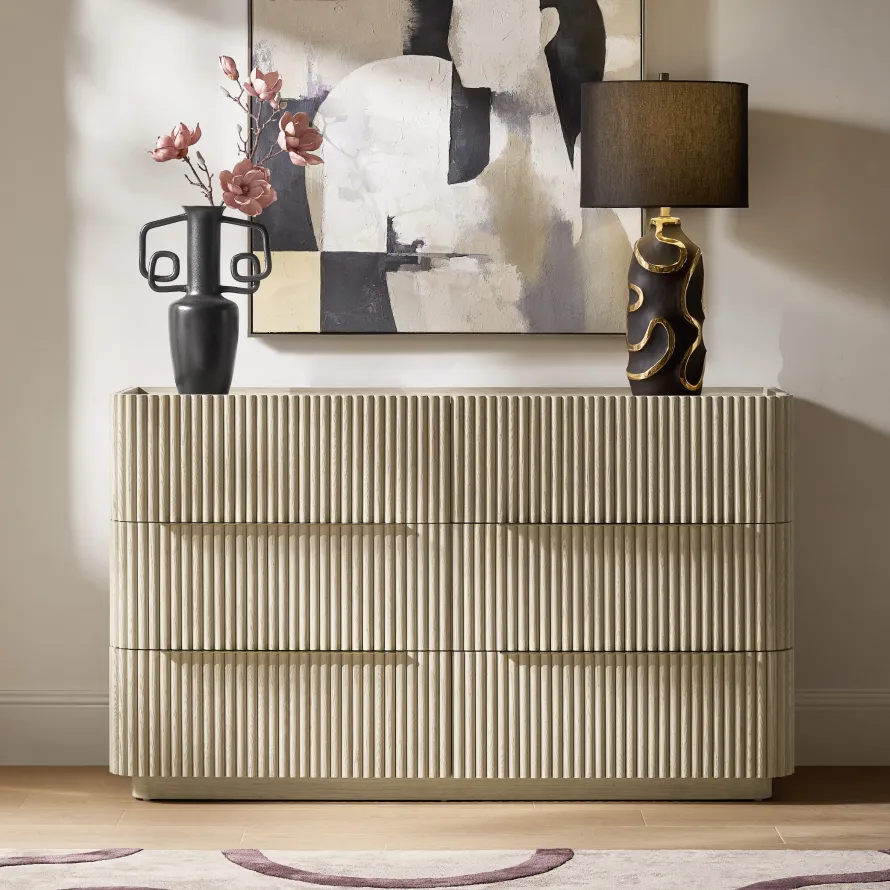 A modern light oak 6-drawer dresser with fluted paneling, topped with a black ceramic table lamp with gold accents, a black twin-handle vase with pink blossoms, and abstract canvas art.
