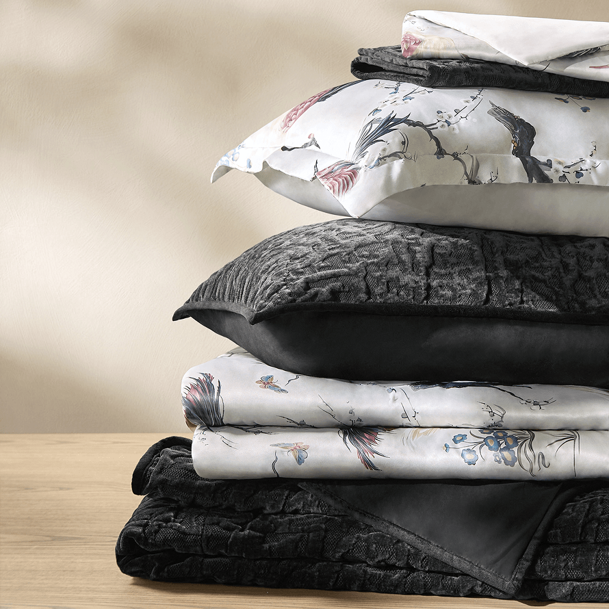 Folded stack of luxury bedding featuring an Asian-inspired floral bird print duvet and shams paired with a textured black velvet quilt and pillows.