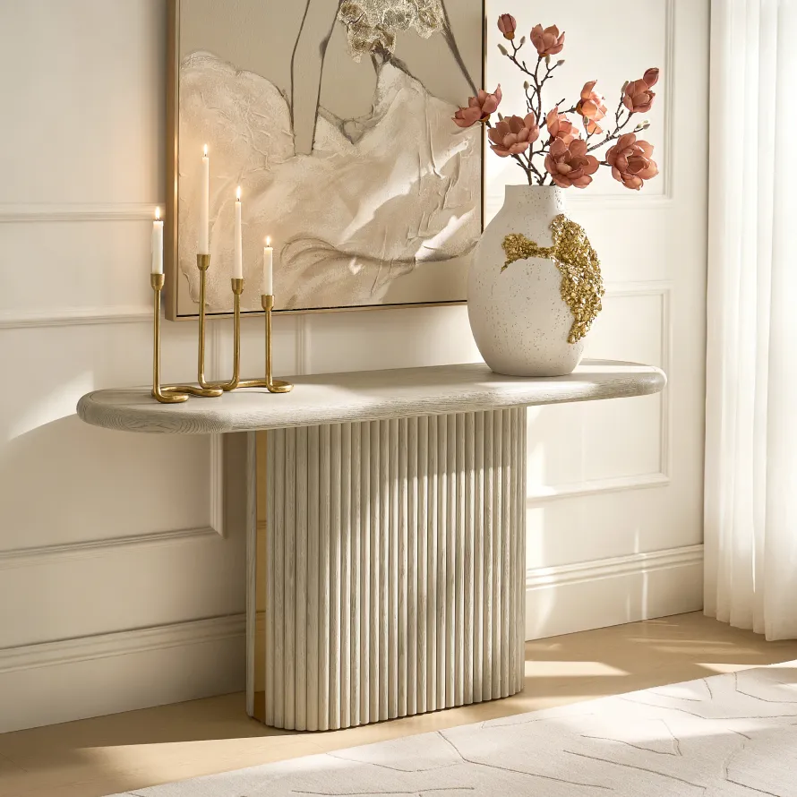 A light oak oval console table with a fluted pedestal base, styled with a gold candelabra, a speckled vase with pink magnolia blossoms, and abstract wall art.